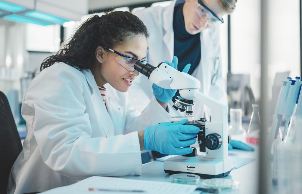 Modern Medical Research Laboratory: Portrait of Two Young Scientists Microscope, Digital Tablet, Doing Sample Analysis, Talking. Diverse Team of Multi-Ethnic Specialists work in Advanced Lab